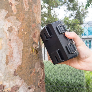 Person holding a surveillance camera device against a tree with a blurred outdoor background