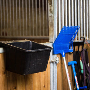 Black feed trough and blue grooming tools against a wooden stable wall.