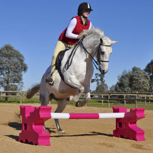 Person riding a white horse jumping over pink and white hurdles in an outdoor equestrian setting.
