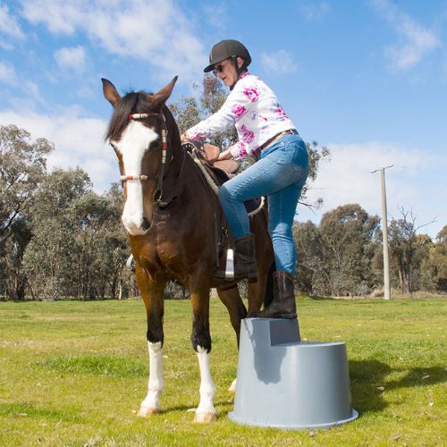 Person getting on a horse using a gray step stool in an outdoor setting