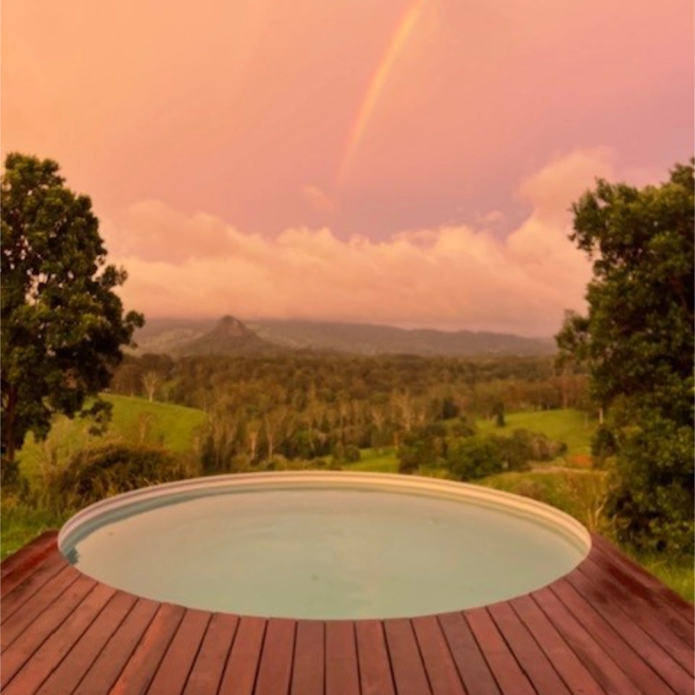 Small above-ground pool on a wooden deck with a scenic view of trees and mountains under a rainbow.