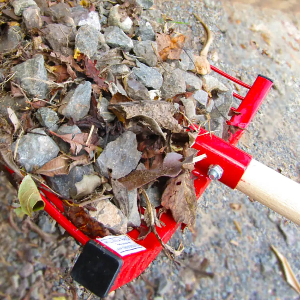 Red garden rake with a wooden handle collecting leaves and small rocks on a driveway.
